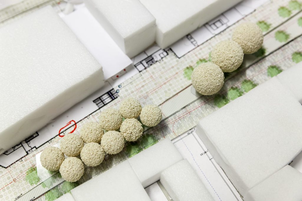 Maqueta arquitectónica en construcción con bloques de espuma y árboles en miniatura sobre planos.