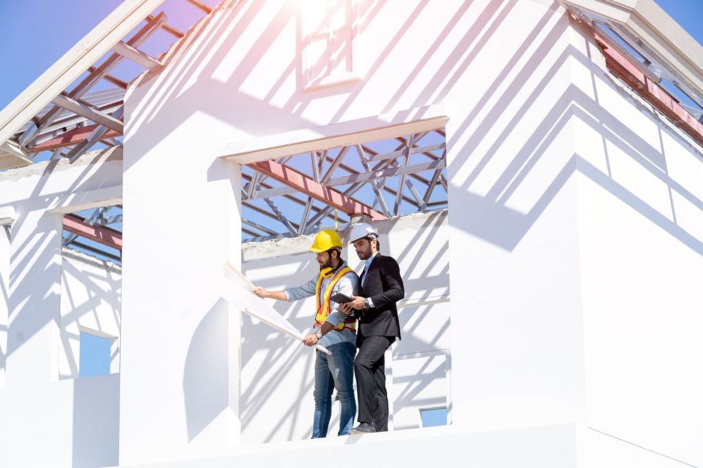 Dos ingenieros examinando planos de construcción en una estructura de casa parcialmente terminada.