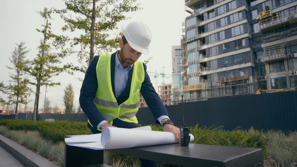 Ingeniero en casco y chaleco reflectante revisando planos en el sitio de construcción.